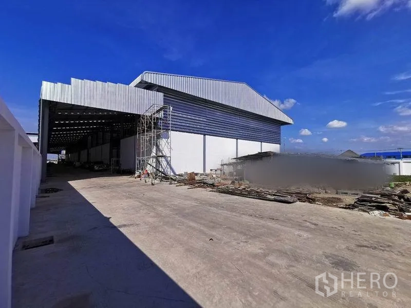 Warehouse for rent in Bang Phli, Samut Prakan - Side elevation of warehouse with canopy, scaffolding and extensive concrete yard under blue sky.