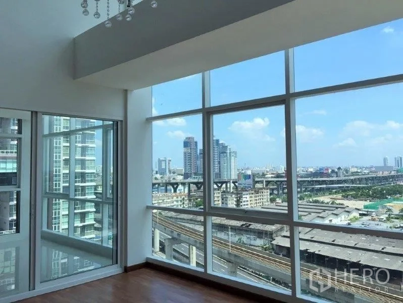 Condominium for sale in Bang Na, Bangkok - Bright corner living area with floor-to-ceiling windows and panoramic Bang Na city views over the elevated rail line.