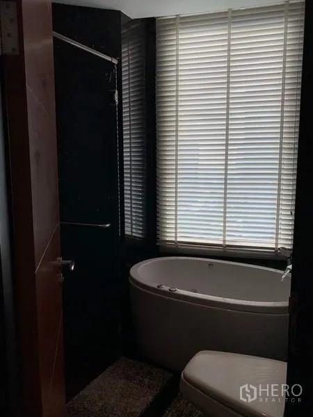 Condominium for rent in Bang Rak, Bangkok - Modern bathroom with an oval bathtub beside a large window with blinds and a separate shower.