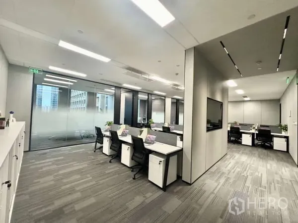 Office Space for rent in Bang Kapi, Bangkok - Corner view of the office showing desks on both sides, a glass wall with city outlook, and an integrated display screen.
