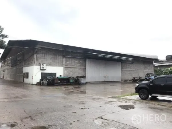 Warehouse for rent in Mueang Pathum Thani, Pathum Thani - Industrial warehouse exterior with large sliding metal doors and a wide concrete yard on a rainy day.