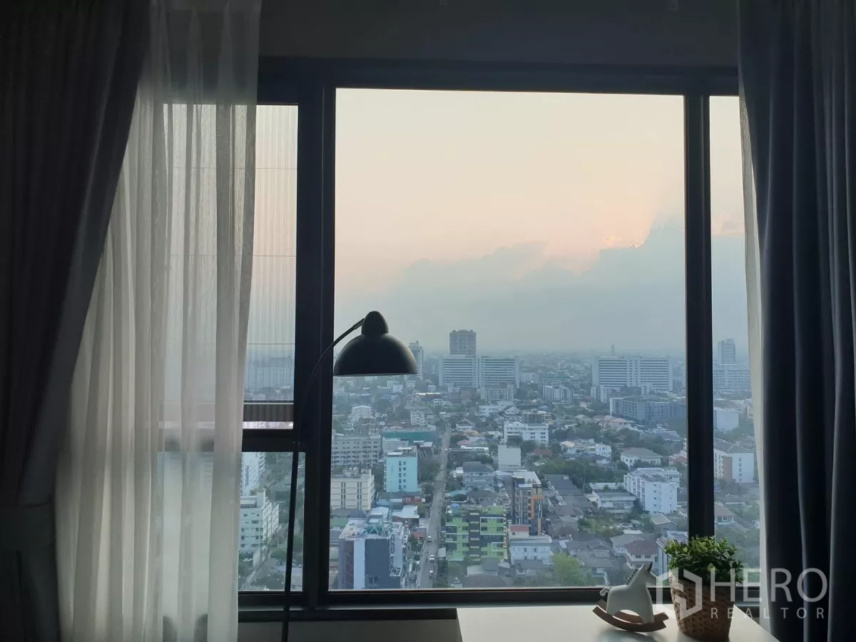 Condominium for rent in Chatuchak, Bangkok - High-floor window view over Chatuchak at sunset with curtains and desk lamp.