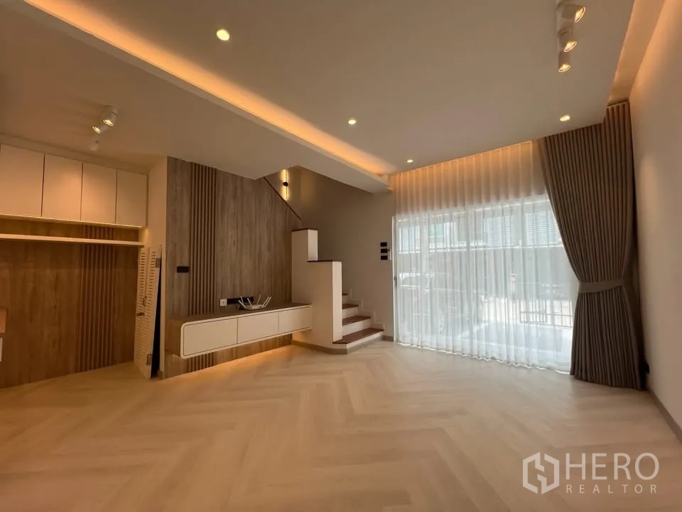 联排别墅 for rent in Prawet, Bangkok - 客厅带楼梯、装饰墙与整面落地窗纱帘。