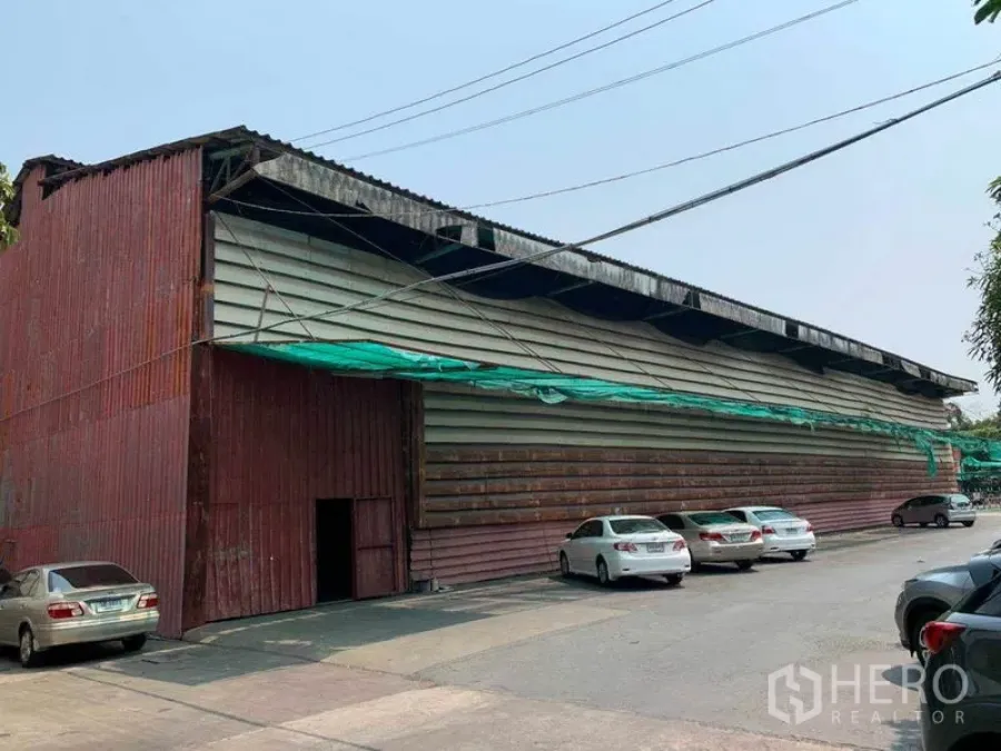 Warehouse for rent in Bang Kho Laem, Bangkok - Street view of a large corrugated‑metal warehouse with front parking and wide driveway.