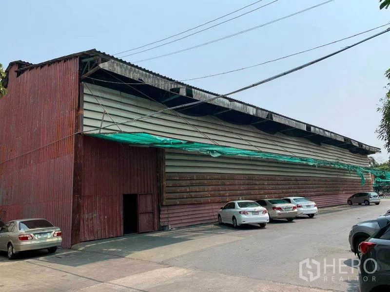 Warehouse for rent in Bang Kho Laem, Bangkok - Street view of a large corrugated‑metal warehouse with front parking and wide driveway.