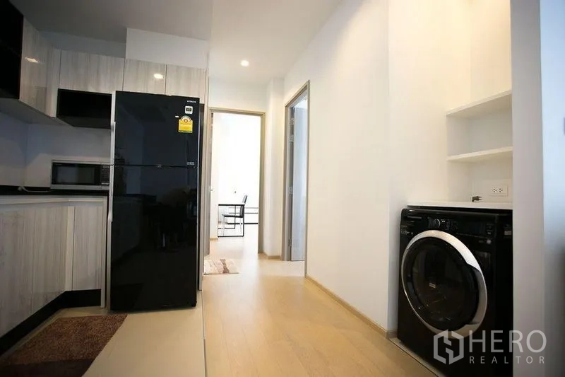 Condominium for rent in Watthana, Bangkok - Hallway view of open kitchen, glossy black fridge and built-in washing machine.