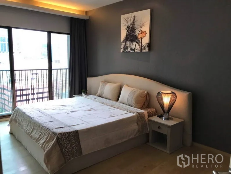 Condominium for rent in Bangkok - Bedroom with king bed, bedside lamp, and floor-to-ceiling windows opening to a balcony with city views.