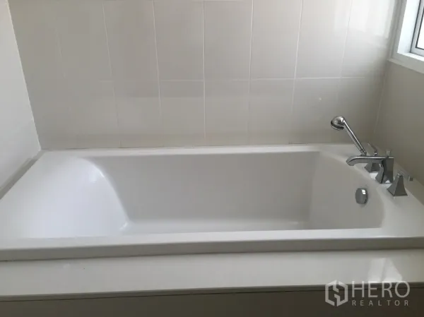Townhouse for rent in Watthana, Bangkok - Clean bathroom featuring a white built-in bathtub with handheld shower by the window.