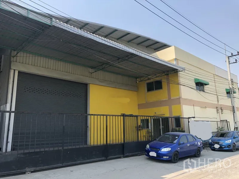 工厂 出租在Pathum Thani - 沿街视角的工厂外立面，金属雨棚、高栅栏大门与路边停车位，位于工业区。