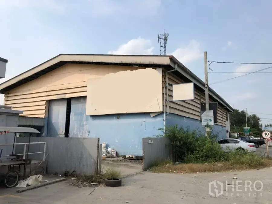 คลังสินค้า ให้เช่าในNonthaburi - ด้านหน้าโกดังหลังคาทรงจั่ว ผนังสีฟ้า ประตูเหล็กและลานจอด ติดถนนสายย่อย