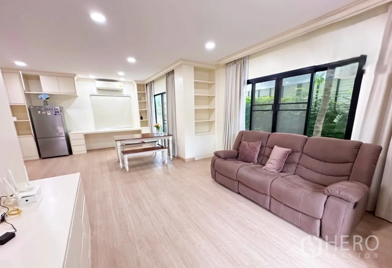 Detached House for sale in Lam Luk Ka, Pathum Thani - Open-plan living area with brown sofa, built-in shelving, dining table and large windows to greenery.