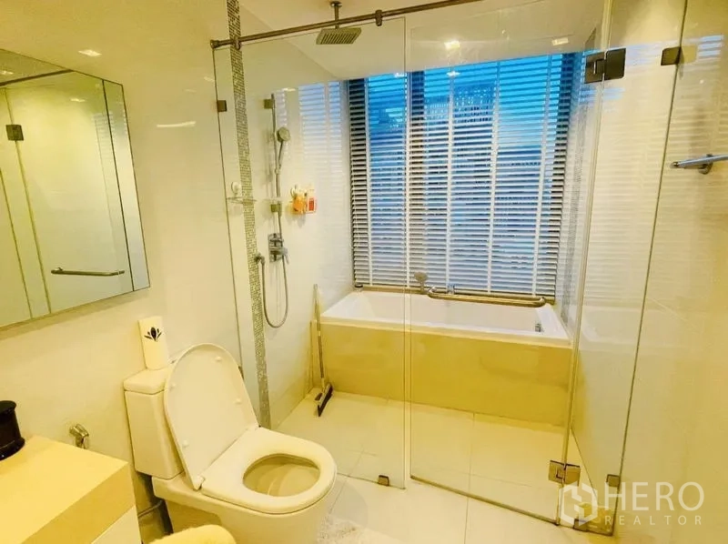 Condominium for rent or sale in Sathon, Bangkok - Modern bathroom with glass shower enclosure, bathtub and window blinds.