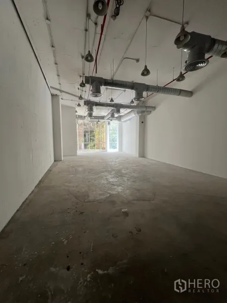 Retail Space for rent - Long open-plan shell retail interior with high ceilings, exposed ductwork and pendant lights, concrete floor, and a bright window at the end.
