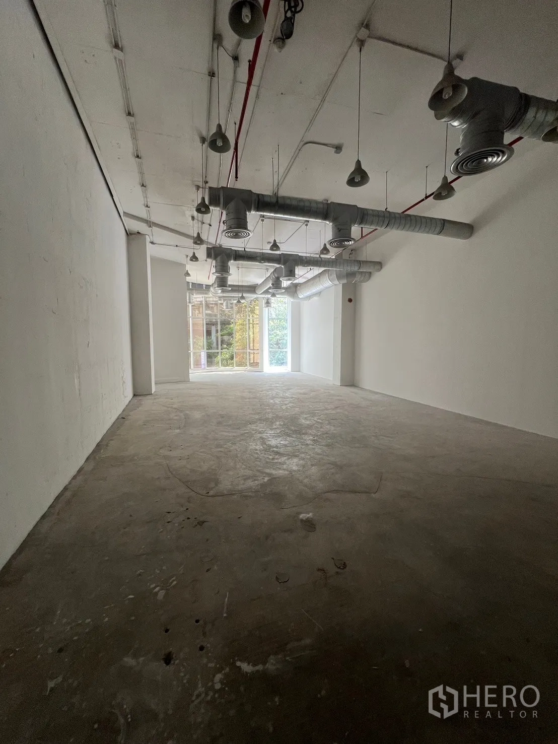 Retail Space for rent - Long open-plan shell retail interior with high ceilings, exposed ductwork and pendant lights, concrete floor, and a bright window at the end.