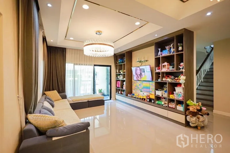 Detached House for rent or sale in Lam Luk Ka, Pathum Thani - Family lounge with built-in TV wall, shelving and chandelier lighting.