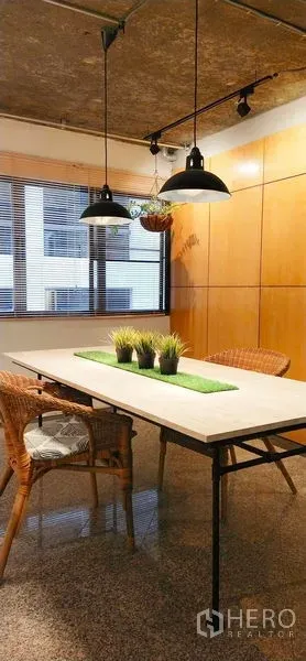 Condominium for sale in Watthana, Bangkok - Bangkok condo dining area with long table, rattan chairs and industrial pendant lights by a window and wood panel wall.