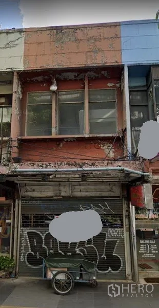 Shophouse for rent in Khlong Toei, Bangkok - Two-storey shophouse exterior with metal roll-up shutter, weathered facade, overhead utility cables and sidewalk frontage on a city street.