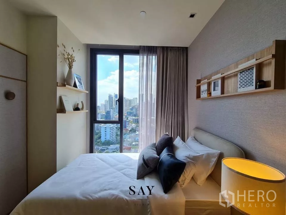 Condominium for rent or sale in Khlong Toei, Bangkok - Cozy bedroom with bed by tall window, floating wall shelves and a clear city skyline view.