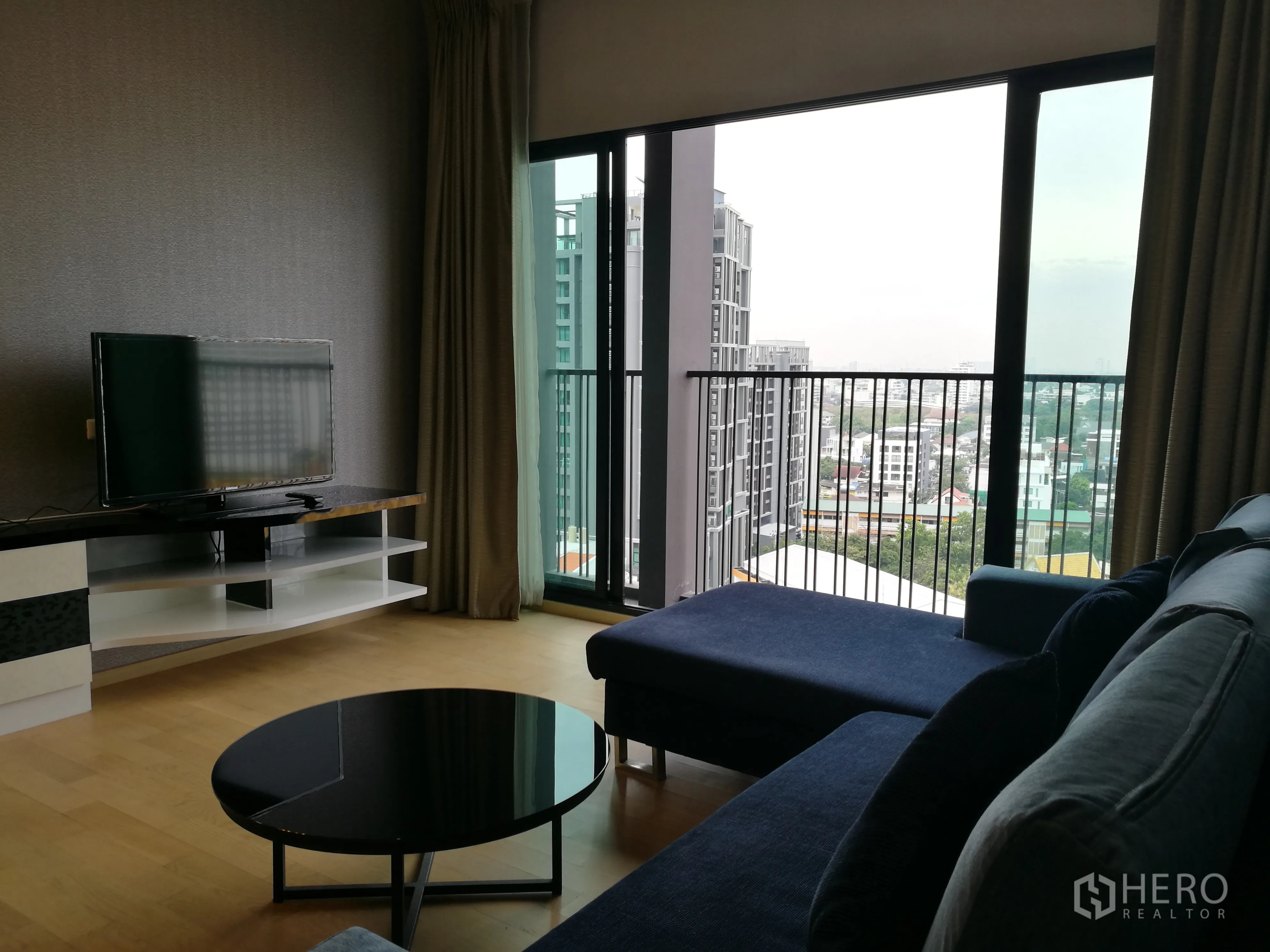 คอนโด for rent in Watthana, Bangkok - ห้องนั่งเล่นพร้อมโซฟาเข้ามุมสีน้ำเงิน ชั้นวางทีวี และประตูกระจกสูงจรดเพดานเปิดออกสู่ระเบียงชมวิวเมือง