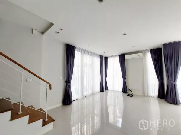 Townhouse for rent in Lat Phrao, Bangkok - Open-plan living area with glossy tile floors, a wooden-step staircase and full-height windows.