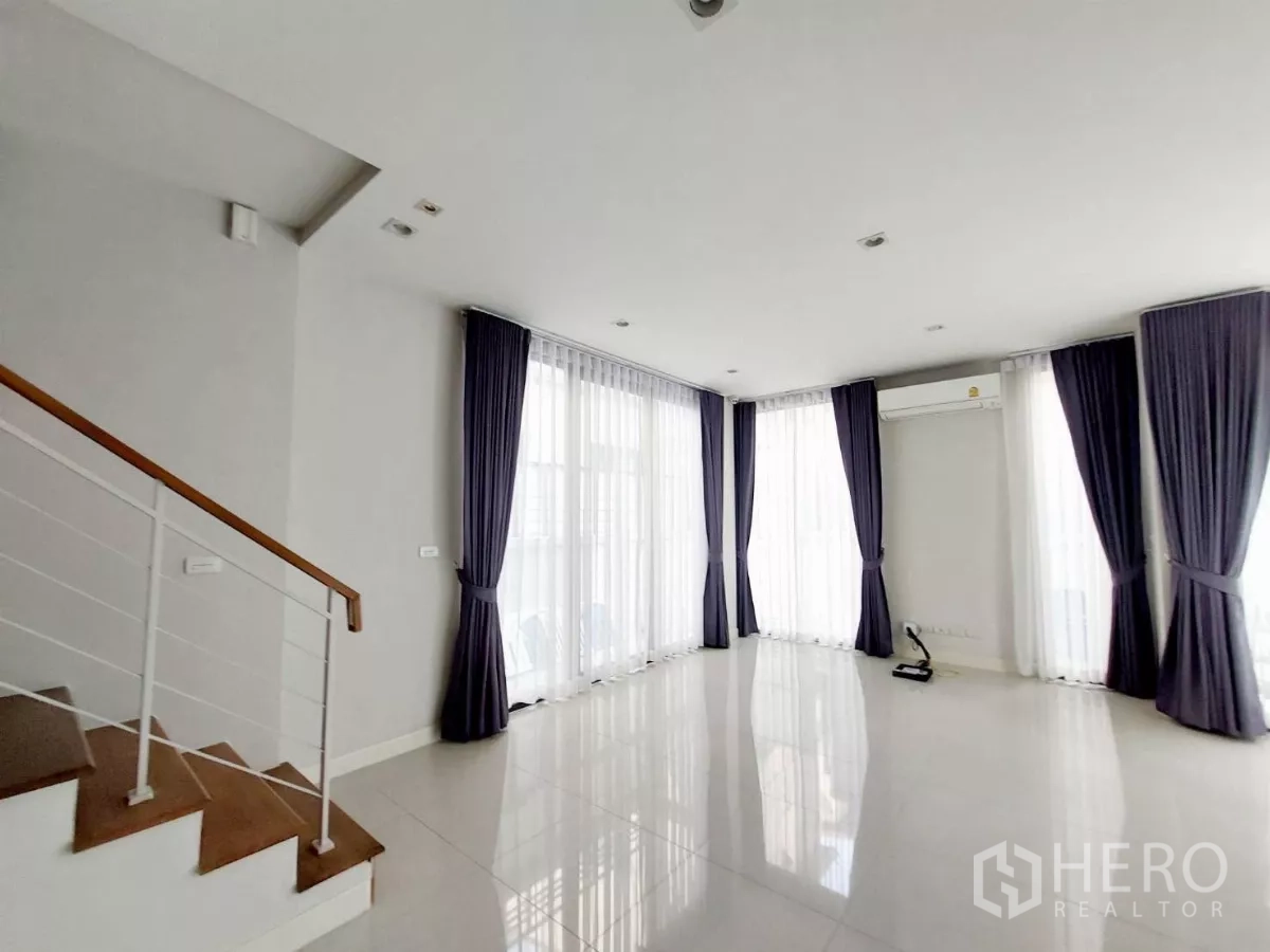 Townhouse for rent in Lat Phrao, Bangkok - Open-plan living area with glossy tile floors, a wooden-step staircase and full-height windows.