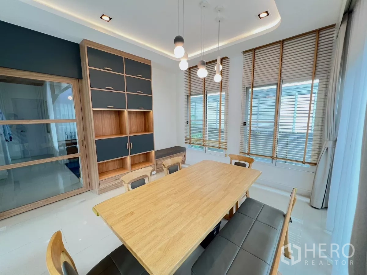 Detached House for rent or sale in Khlong Luang, Pathum Thani - Dining area with wooden table, pendant lights and corner windows with blinds.