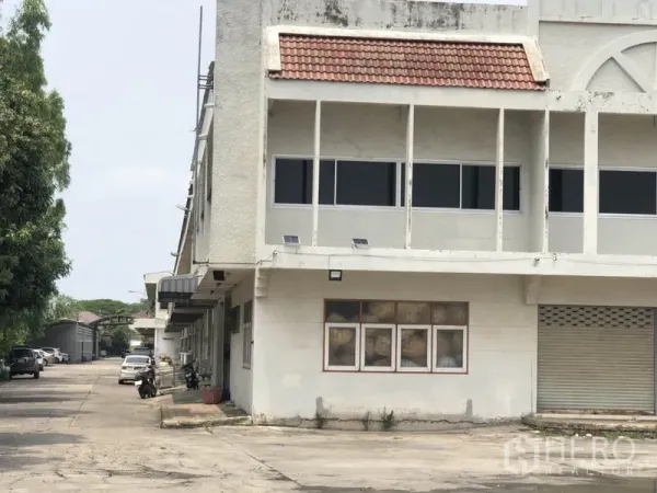 货栈 出租在Bang Kapi, Bangkok - 仓库转角特写，带遮棚走道、窗户与卷闸门，旁为绿树成荫的车道。