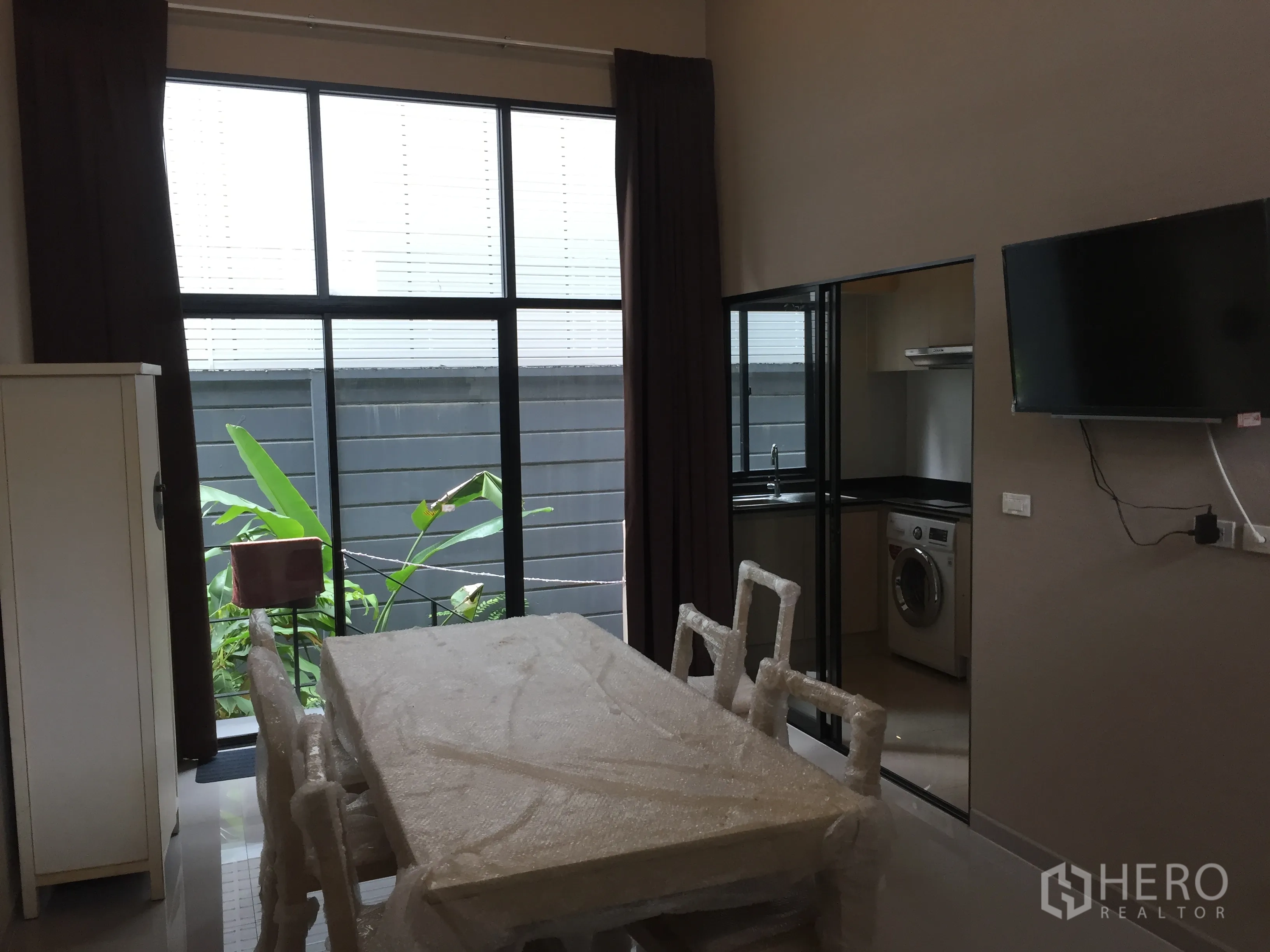 Townhouse for rent in Suan Luang, Bangkok - Dining area with floor‑to‑ceiling windows opening to a small garden, TV on wall and sliding door to kitchen.