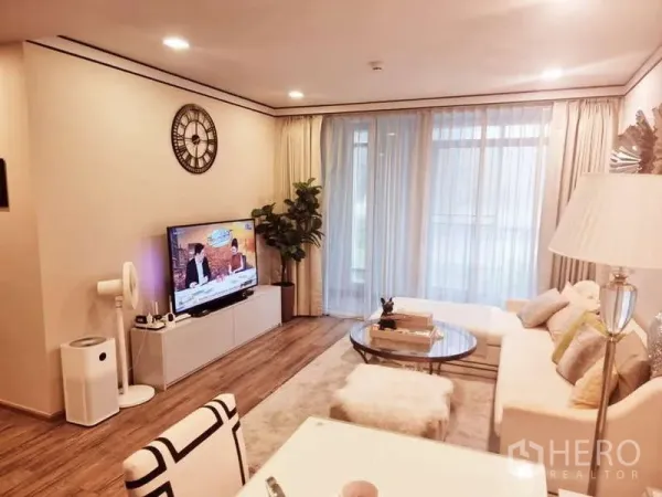 Condominium for sale in Bang Rak, Bangkok - Bright living room with white sofa, round glass coffee table, TV console, indoor plant and full-height sheer curtains over wood floors.