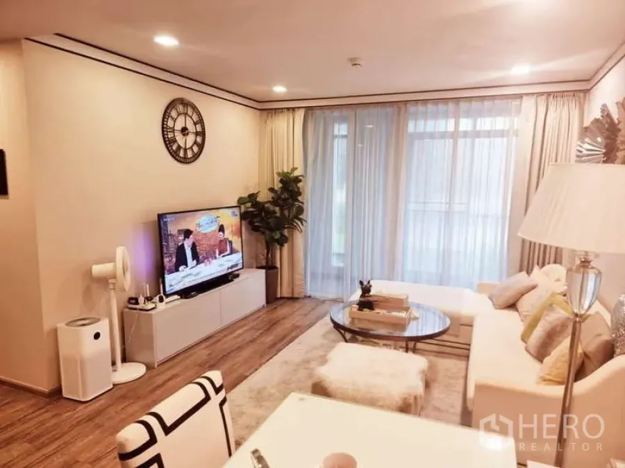 Condominium for sale in Bang Rak, Bangkok - Bright living room with white sofa, round glass coffee table, TV console, indoor plant and full-height sheer curtains over wood floors.