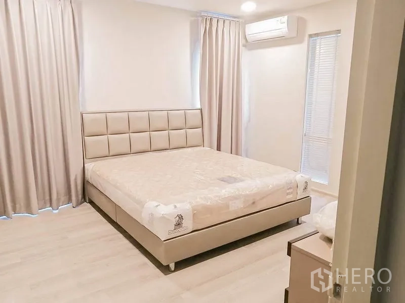 Townhouse for rent in Prawet, Bangkok - Primary bedroom with upholstered bed, soft curtains, air conditioning and warm wood flooring.