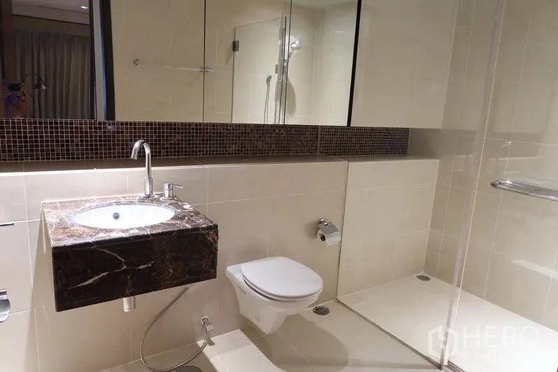 Condominium for rent in Khlong Toei, Bangkok - Second bathroom with walk-in glass shower, wall-mounted toilet and marble sink counter.