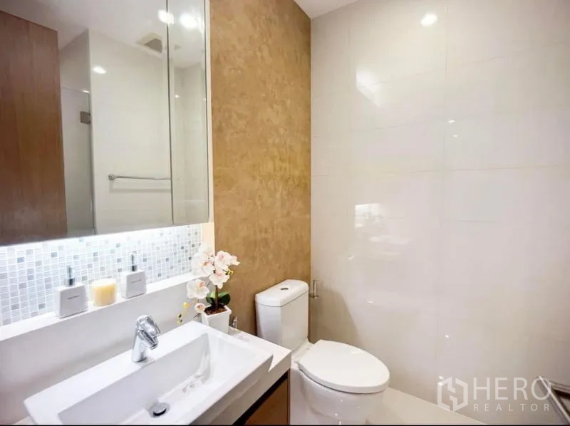 Condominium for rent in Khlong Toei, Bangkok - Contemporary bathroom with wall-mounted sink, toilet and backlit mirror in neutral tones.