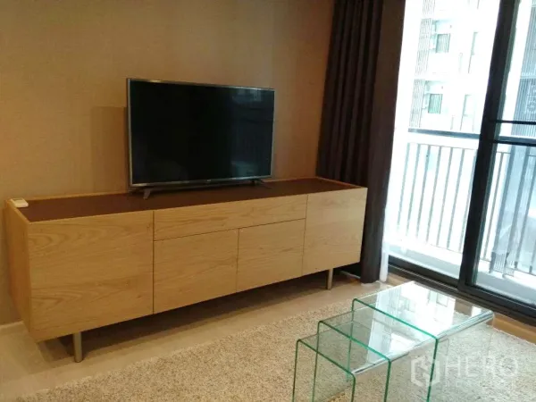 Condominium for rent in Khlong Toei, Bangkok - Living area with wood TV console, glass nesting tables, and sliding door opening to the balcony.