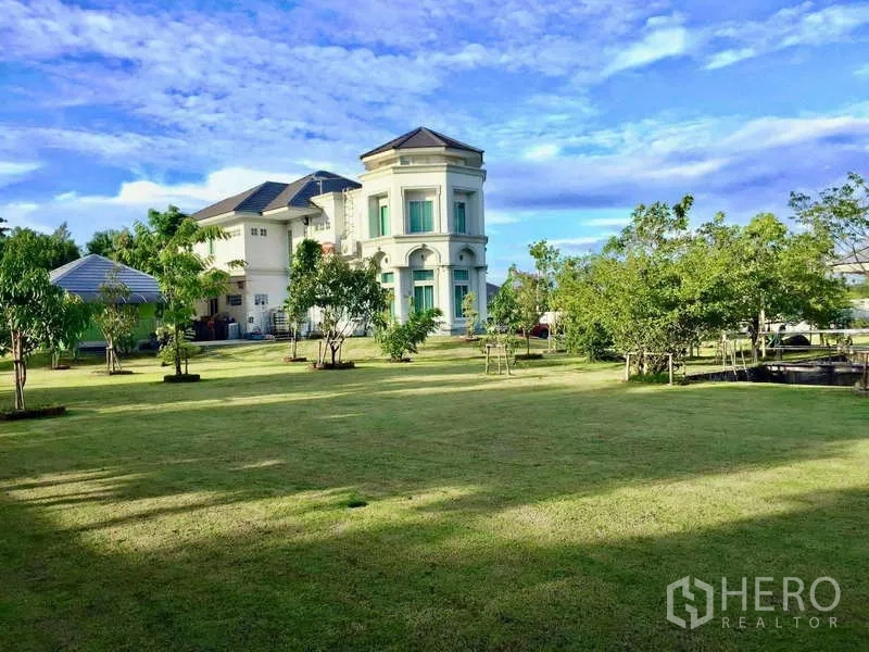 Detached House for sale in Khlong Luang, Pathum Thani - Vast open lawn with trees leading to a white two-storey residence and outbuildings.