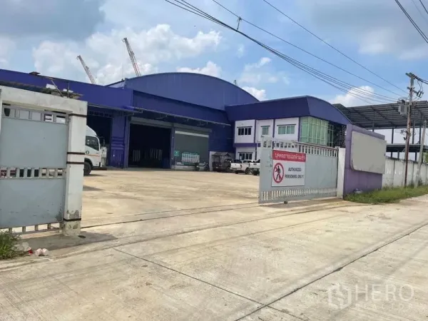 Warehouse for sale in Lam Luk Ka, Pathum Thani - Wide driveway leading to loading bays and main warehouse facade with sliding gates and ample truck space.
