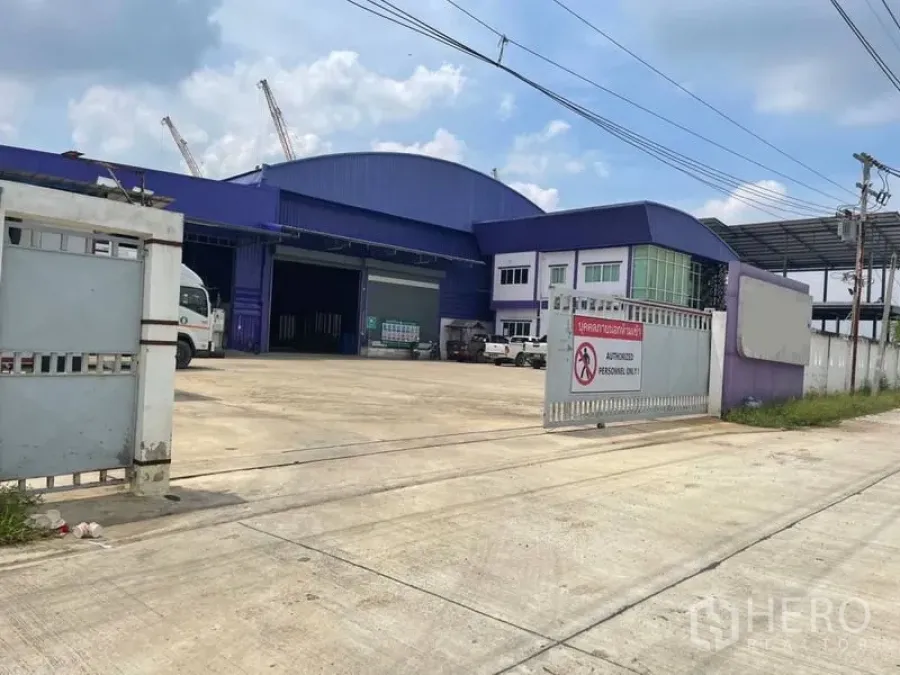 Warehouse for sale in Lam Luk Ka, Pathum Thani - Wide driveway leading to loading bays and main warehouse facade with sliding gates and ample truck space.
