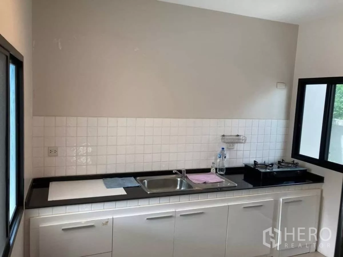 Detached House for rent in Mueang Nonthaburi, Nonthaburi - Closed kitchen with white cabinetry, sink and gas hob next to corner windows.