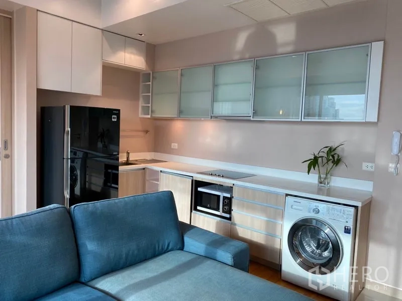 Condominium for rent or sale in Yan Nawa, Bangkok - Open-plan kitchen with glass-front cabinets, electric hob, microwave, fridge and washing machine beside a blue sofa on wood floors.
