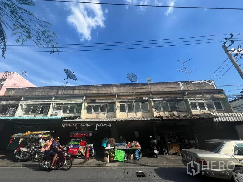 ที่ดิน ให้เช่าหรือขายในPhra Khanong, Bangkok - มุมมองด้านหน้าของตึกแถวเก่าแก่ มีแผงตลาดและรถจักรยานยนต์จอดริมถนน.