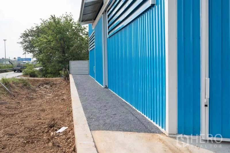 Warehouse for rent in Khlong Luang, Pathum Thani - Gravel path along the blue facade beside the roadway under awnings and vents.