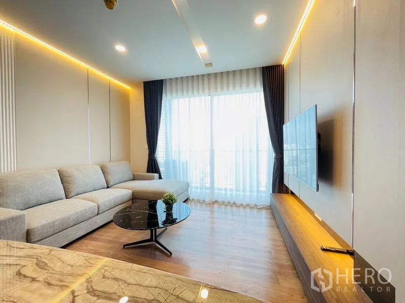 Condominium for rent in Khlong Toei, Bangkok - Bright living room with an L-shaped grey sofa, floor-to-ceiling windows, warm LED lighting and a wall-mounted TV in a modern Bangkok condo near BTS Thonglor.
