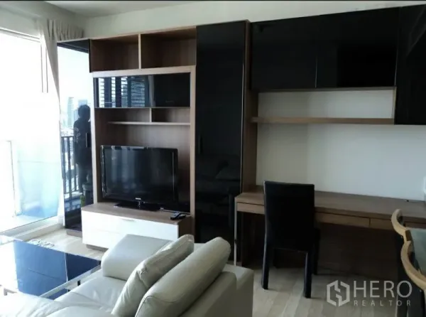 Condominium for rent in Khlong Toei, Bangkok - Living area with TV console, built-in shelving, work desk, and leather sofa beside the balcony.
