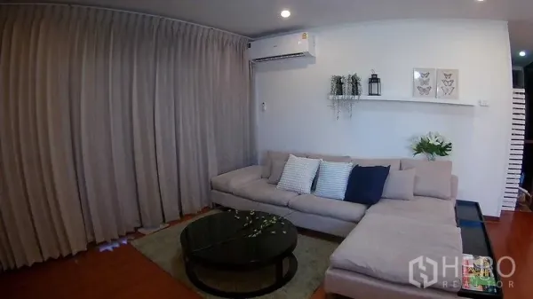 Condominium for rent or sale in Din Daeng, Bangkok - Bright condo living room in Din Daeng, Bangkok with L-shaped sofa, wall-shelf decor, air-conditioner and wood flooring.