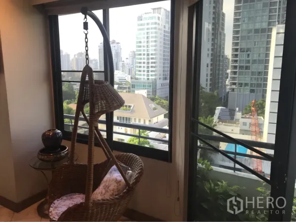 Condominium for rent or sale in Watthana, Bangkok - Corner nook with a rattan hanging chair beside wide windows overlooking the city skyline and greenery.