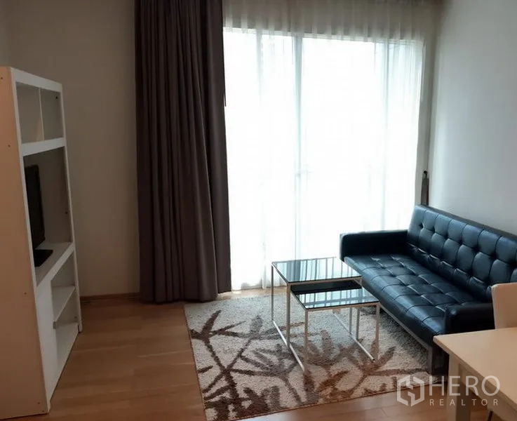 Condominium for rent in Khlong Toei, Bangkok - Bright living room with a black leather sofa, glass coffee tables, TV shelf and floor-to-ceiling window with curtains.