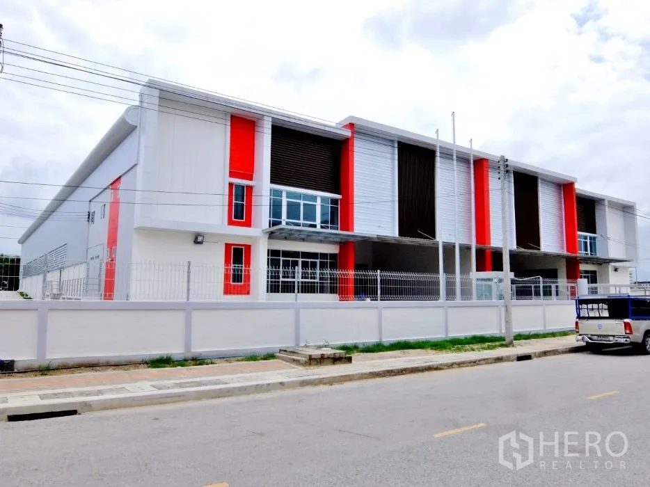 Factory for rent in Bang Sao Thong, Samut Prakan - Front elevation of the industrial building with red accents, fence and driveway.