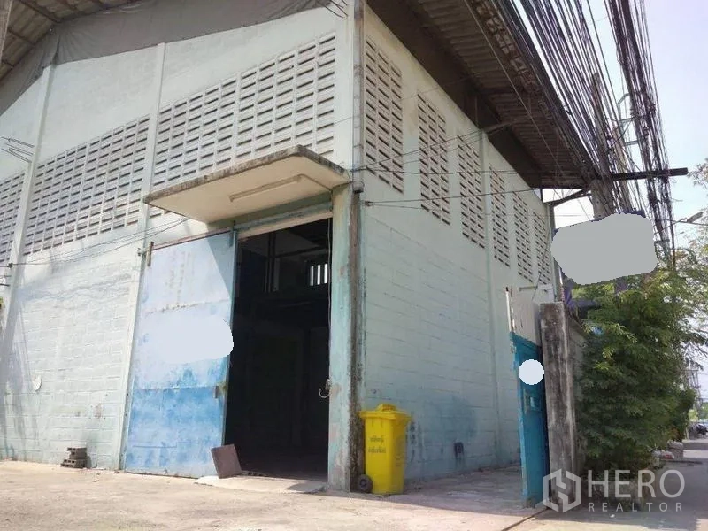 Warehouse for rent in Mueang Nonthaburi, Nonthaburi - Corner warehouse facade with tall sliding door under a small canopy and utility lines along the street.