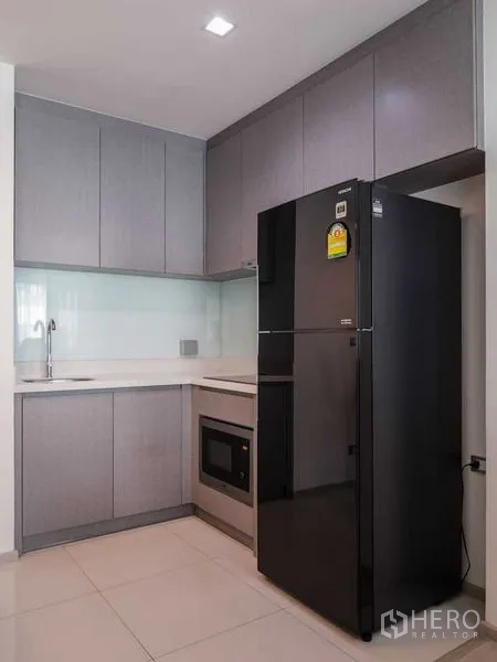 Condominium for rent in Khlong Toei, Bangkok - Compact L-shaped kitchen with grey cabinets, black refrigerator, built-in oven, and sink.