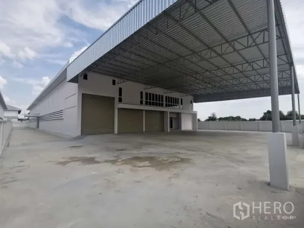 Factory for rent or sale in Bang Bo, Samut Prakan - Side view of factory showing wide driveway and covered loading area with multiple roller shutters.